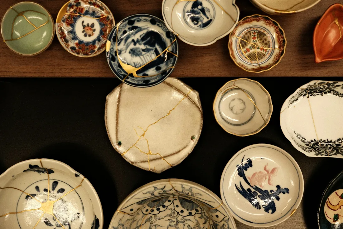 Conjunto de platos y cuencos de cerámica reparados con kintsugi, con las junturas marcadas en dorado, vistos desde arriba sobre una superficie oscura y una tabla de madera.
